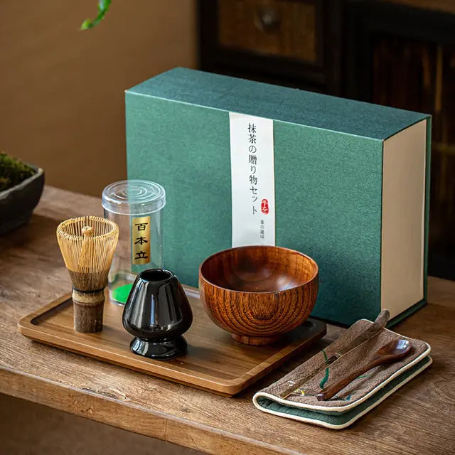 Matcha set with wooden bowl, whisk, and other tools on a wooden tray in front of a green box.